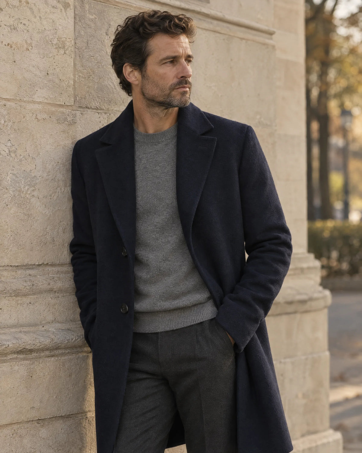 A man in a navy overcoat and grey knit standing against a limestone wall